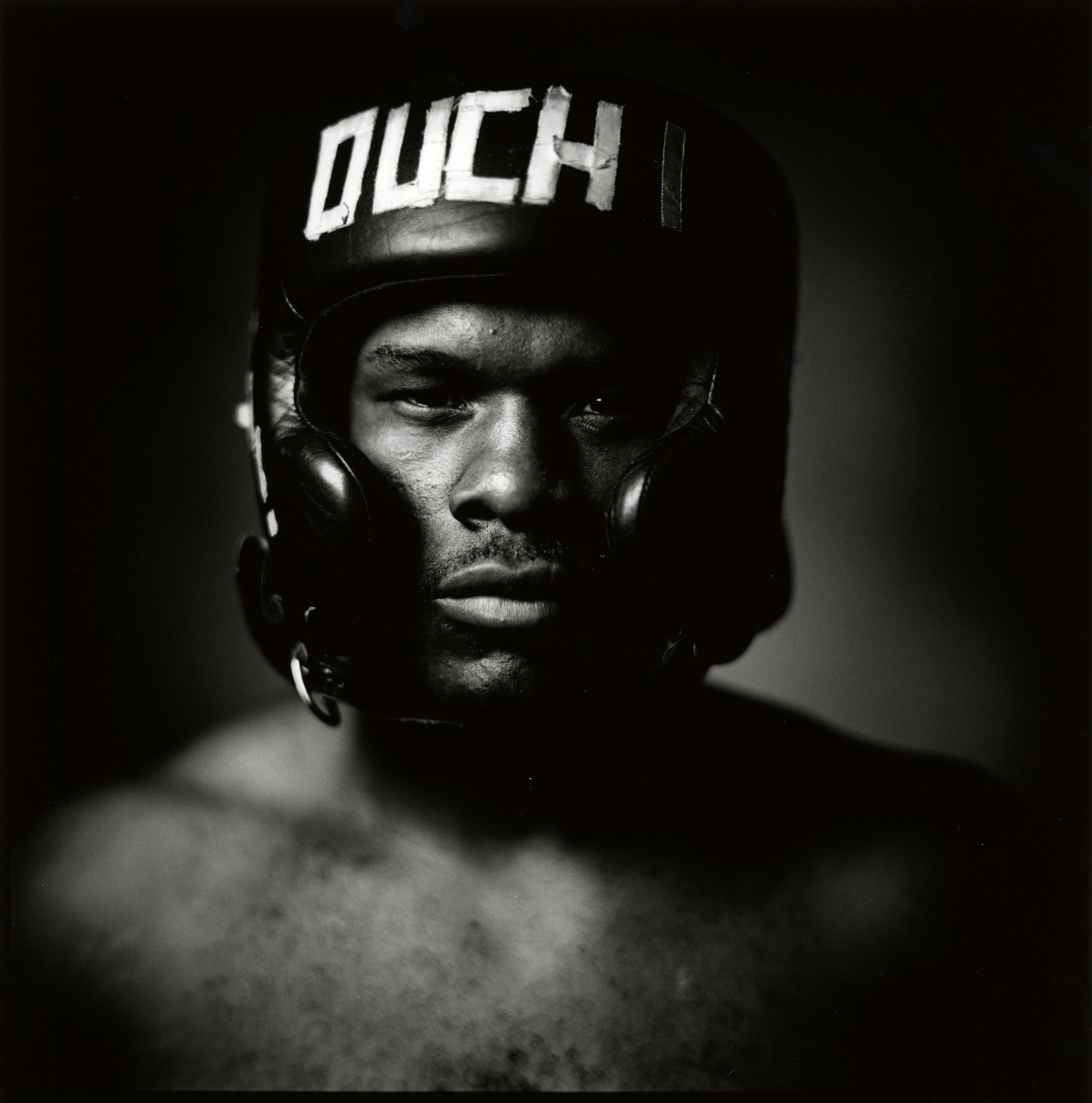 Close-up of a strong-featured adult male wearing a black boxing helmet with the word 'OUCH' in white lettering, dramatic lighting.