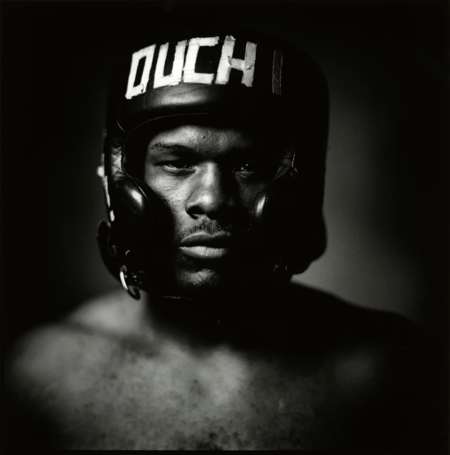 Close-up of a strong-featured adult male wearing a black boxing helmet with the word 'OUCH' in white lettering, dramatic lighting.