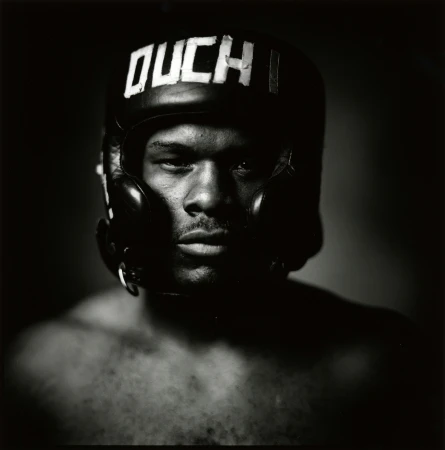 Close-up of a strong-featured adult male wearing a black boxing helmet with the word 'OUCH' in white lettering, dramatic lighting.