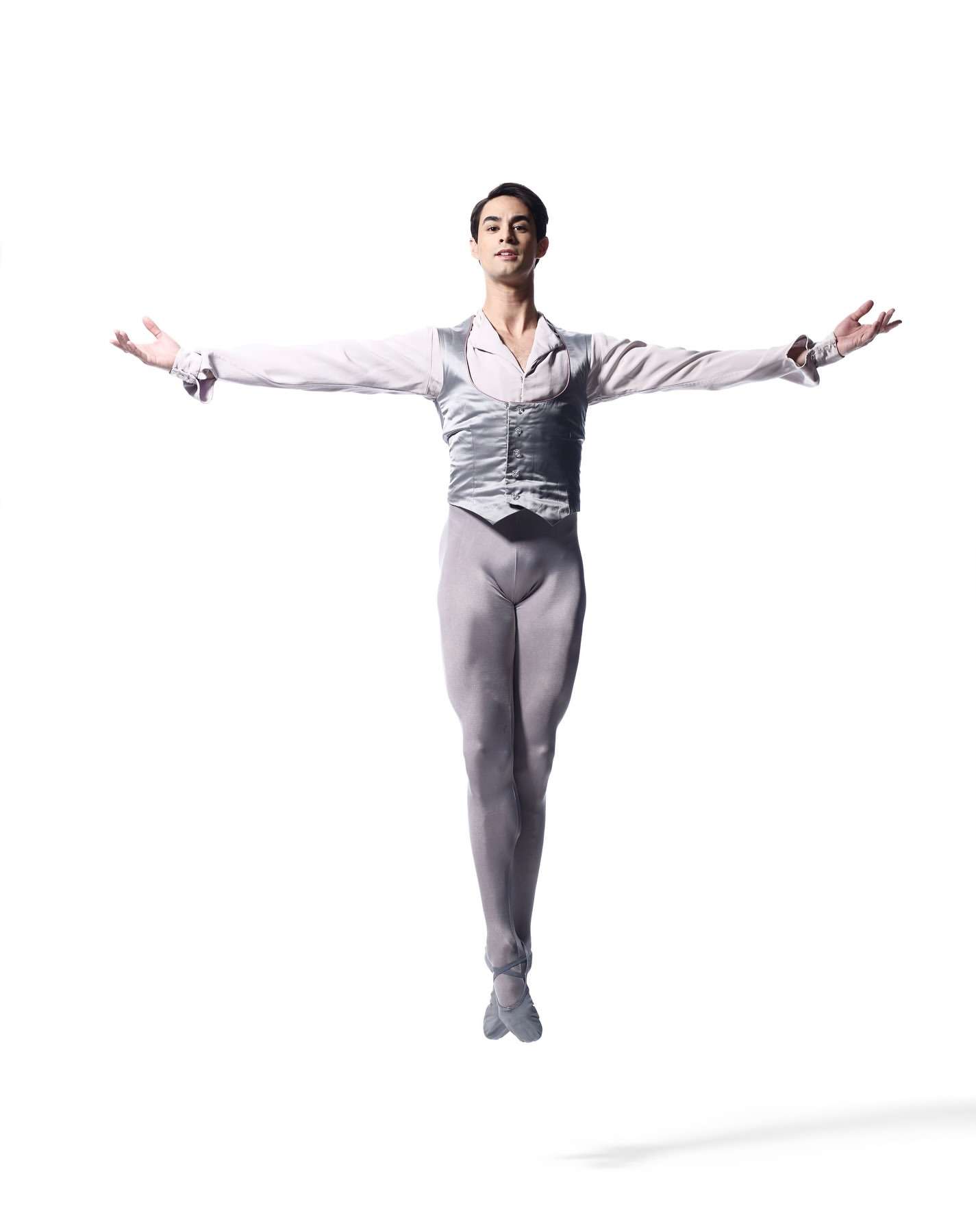Ballet dancer in grey costume leaps in a studio against a white backdrop with outstretched arms and a shadow beneath him.