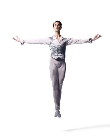 Ballet dancer in grey costume leaps in a studio against a white backdrop with outstretched arms and a shadow beneath him.