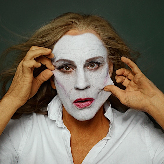 Dramatic portrait of an adult with pale face paint pulling at skin, wearing a white shirt against a dark olive background in studio.