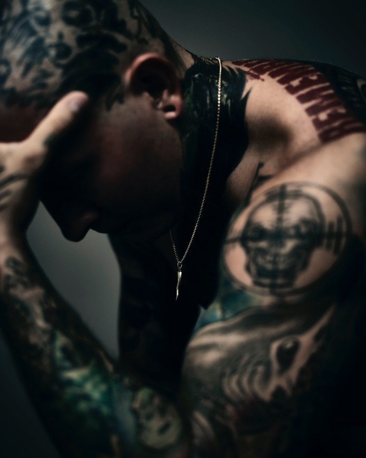 A man with tattoos on his head, arms, and torso has his head in his hands and wears a gold necklace with a pendant.