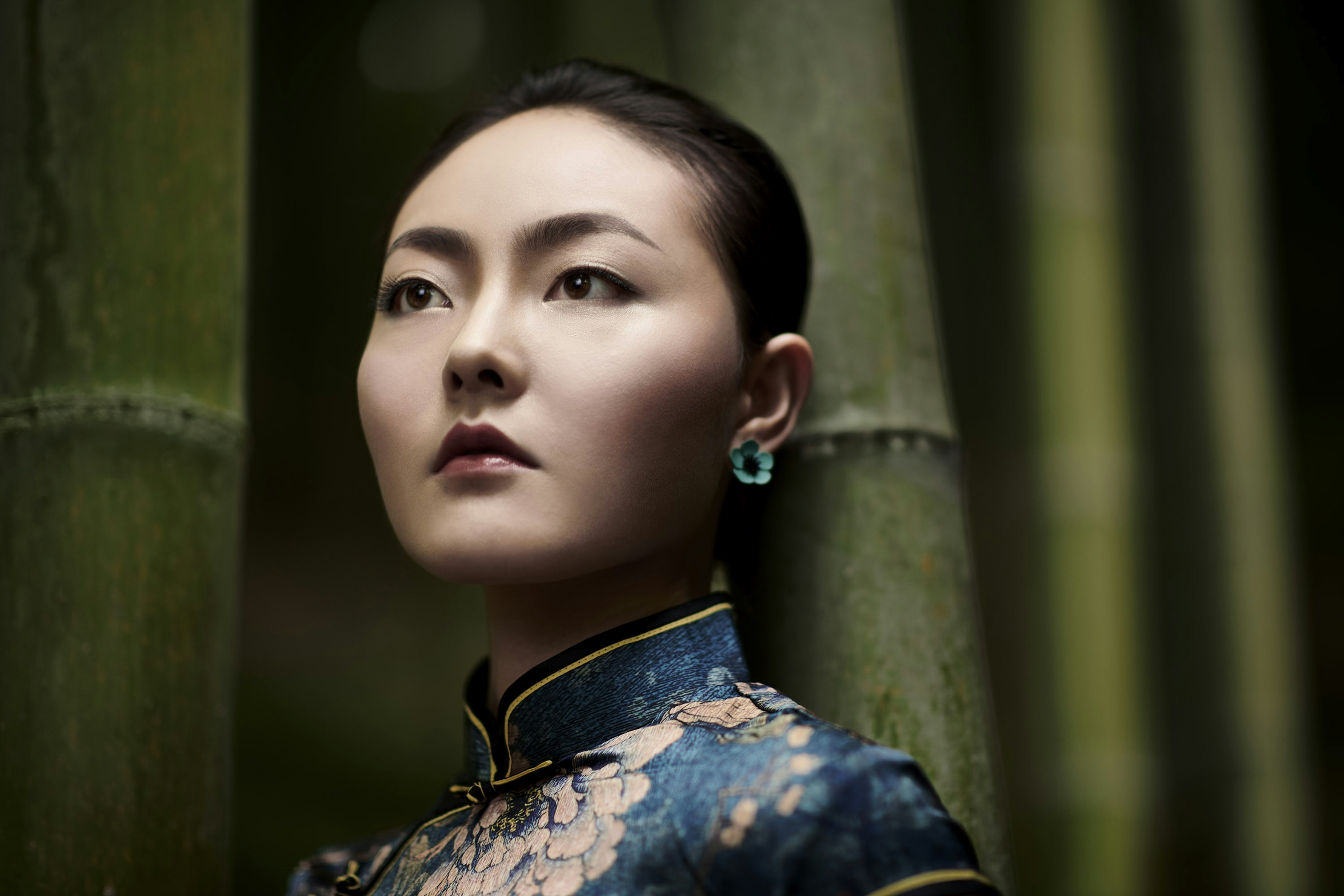 Portrait of an Asian woman with short dark hair in a floral qipao dress standing amongst bamboo stalks, looking off camera.
