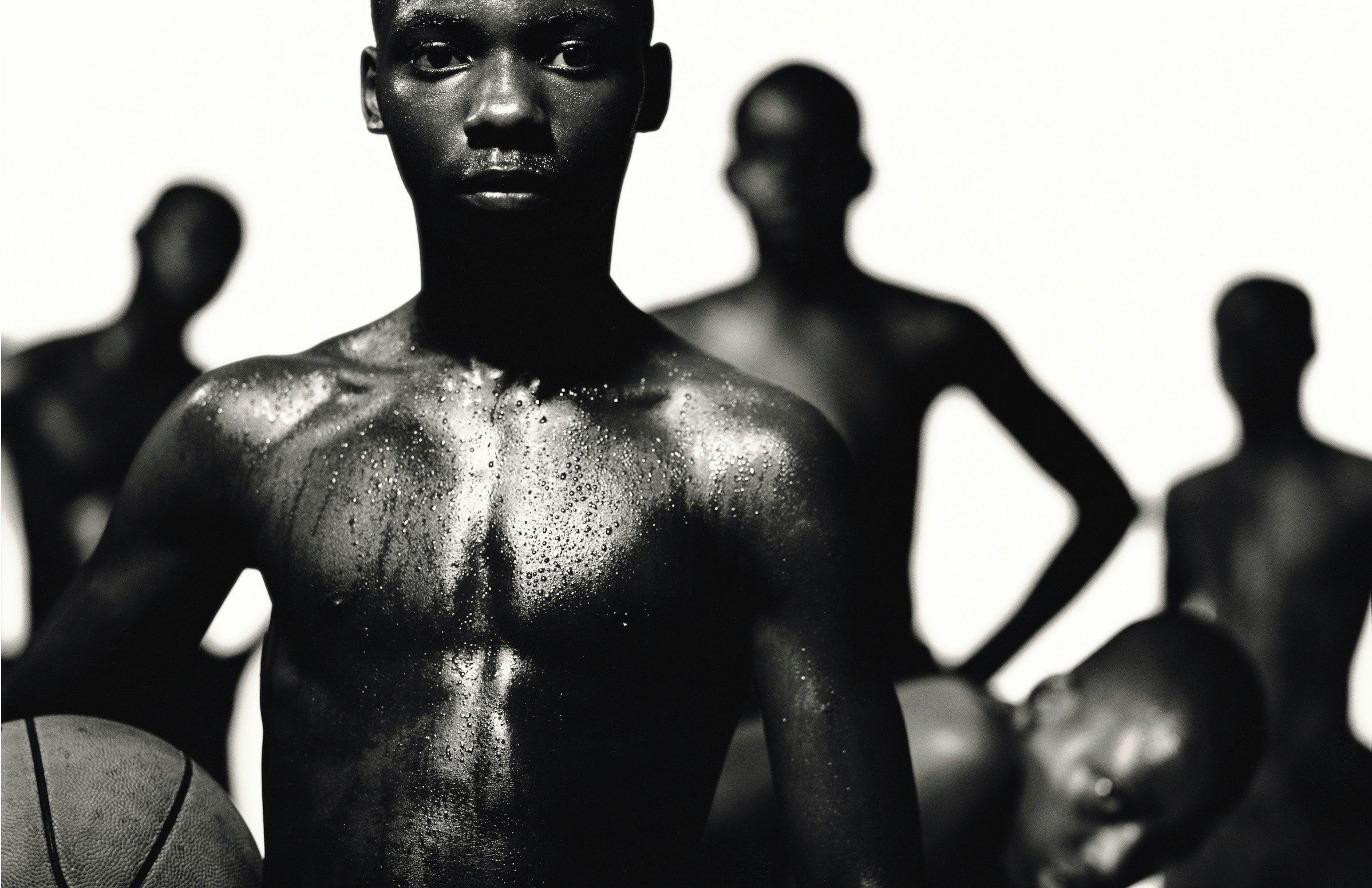 Black and white portrait of a shirtless young Black man holding a basketball, with blurry figures in the background.