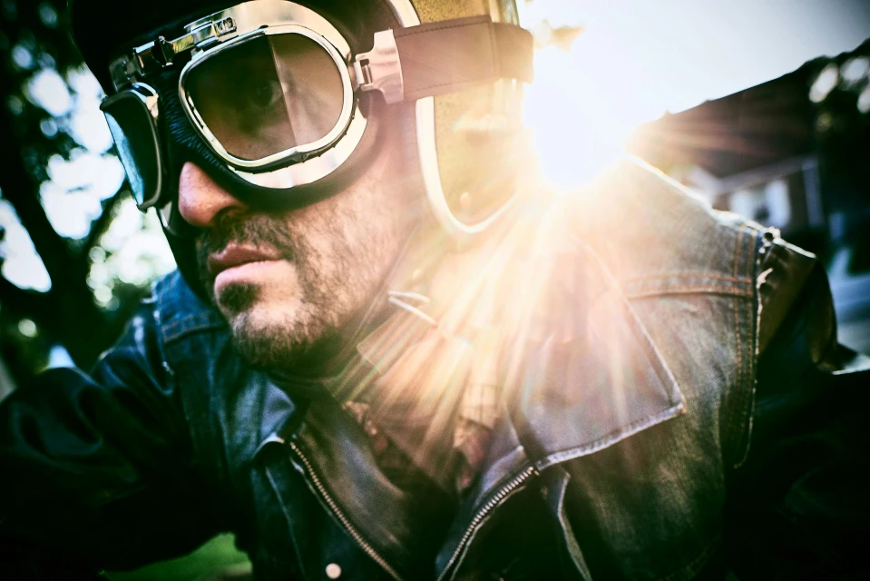 Close-up of adult male wearing a helmet and goggles outdoors with bright sunlight, leather jacket, and a denim vest.
