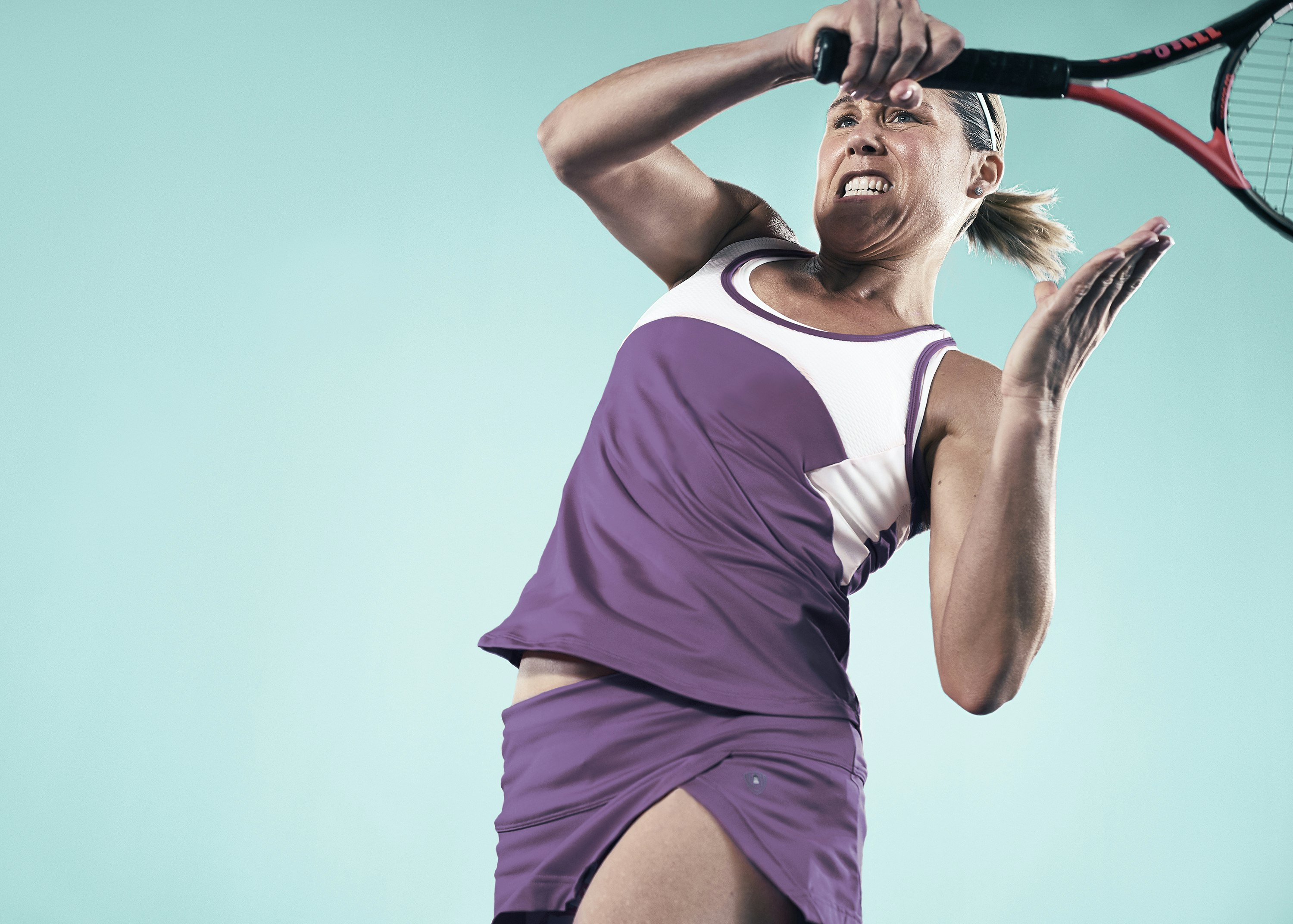 Female tennis player with aggressive facial expression swings racket in a purple athletic outfit against a blue background in a low-angle shot.