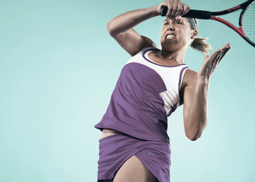 Female tennis player with aggressive facial expression swings racket in a purple athletic outfit against a blue background in a low-angle shot.