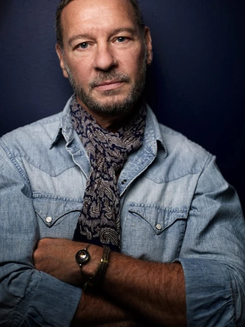 Portrait of a mature adult man with a paisley scarf, wearing a denim shirt with arms crossed against a dark navy blue backdrop.