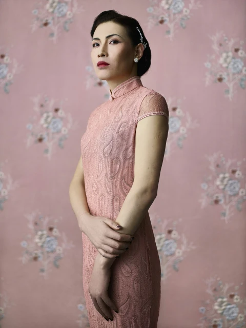 Portrait of an Asian woman in a pink lace dress against a floral backdrop. She poses with hands crossed, side view, wearing earrings.