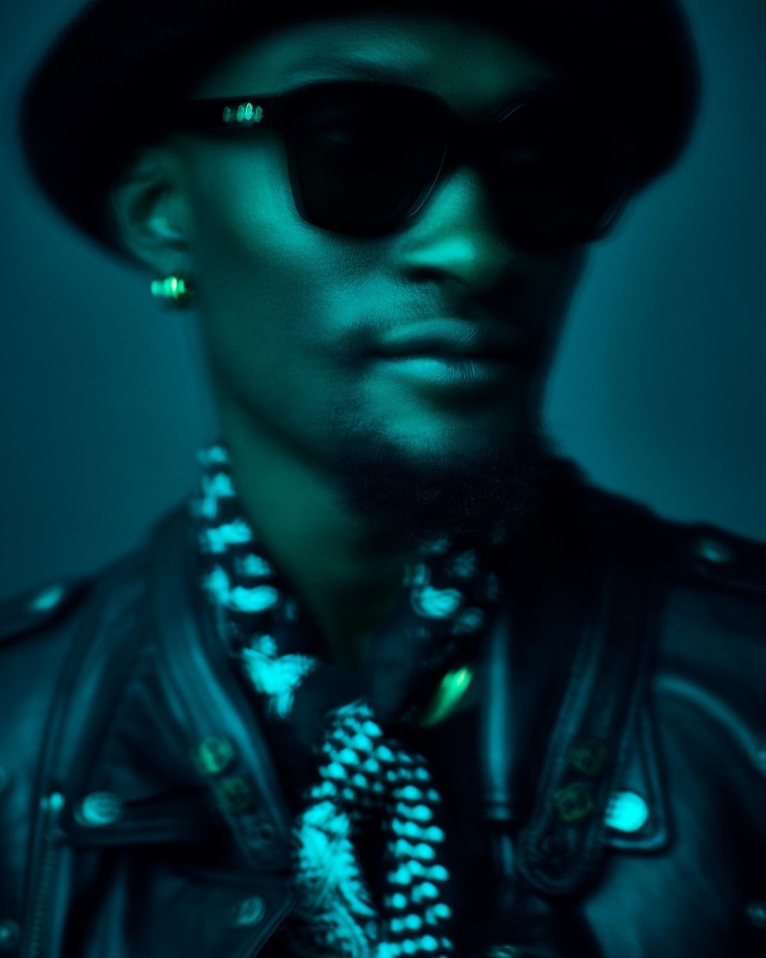 Close-up portrait of a stylish adult male with dark skin in sunglasses, hat, and jacket, against a teal background with a slight blur effect.