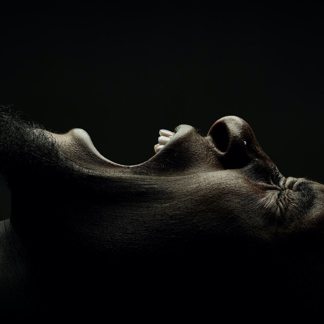 Close-up studio shot of an adult with a dark complexion. The subject's mouth is open, and the chin is tilted upwards against a stark black background.