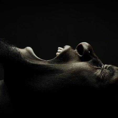 Close-up studio shot of an adult with a dark complexion. The subject's mouth is open, and the chin is tilted upwards against a stark black background.