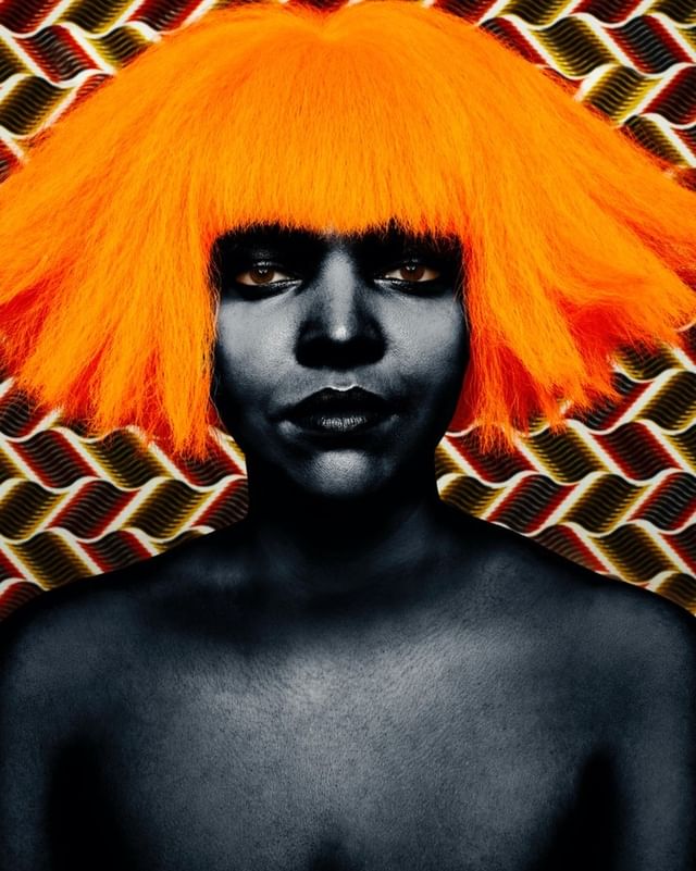 Dramatic portrait of a black-skinned woman with a vibrant orange wig, set against a patterned backdrop, creating a striking contrast.