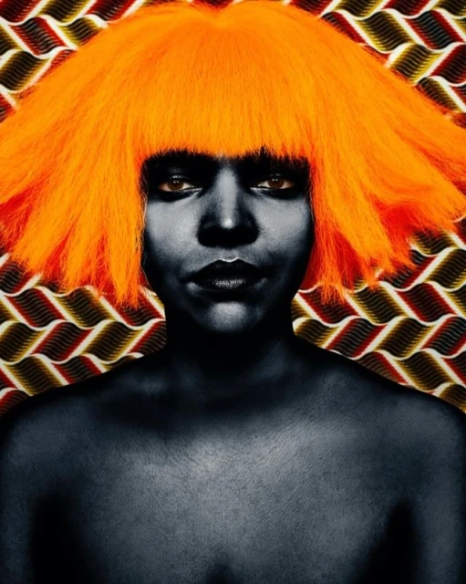 Dramatic portrait of a black-skinned woman with a vibrant orange wig, set against a patterned backdrop, creating a striking contrast.