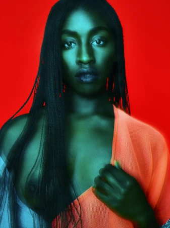 Portrait of a Black woman with long microbraids wearing an orange mesh garment against a vibrant red background.
