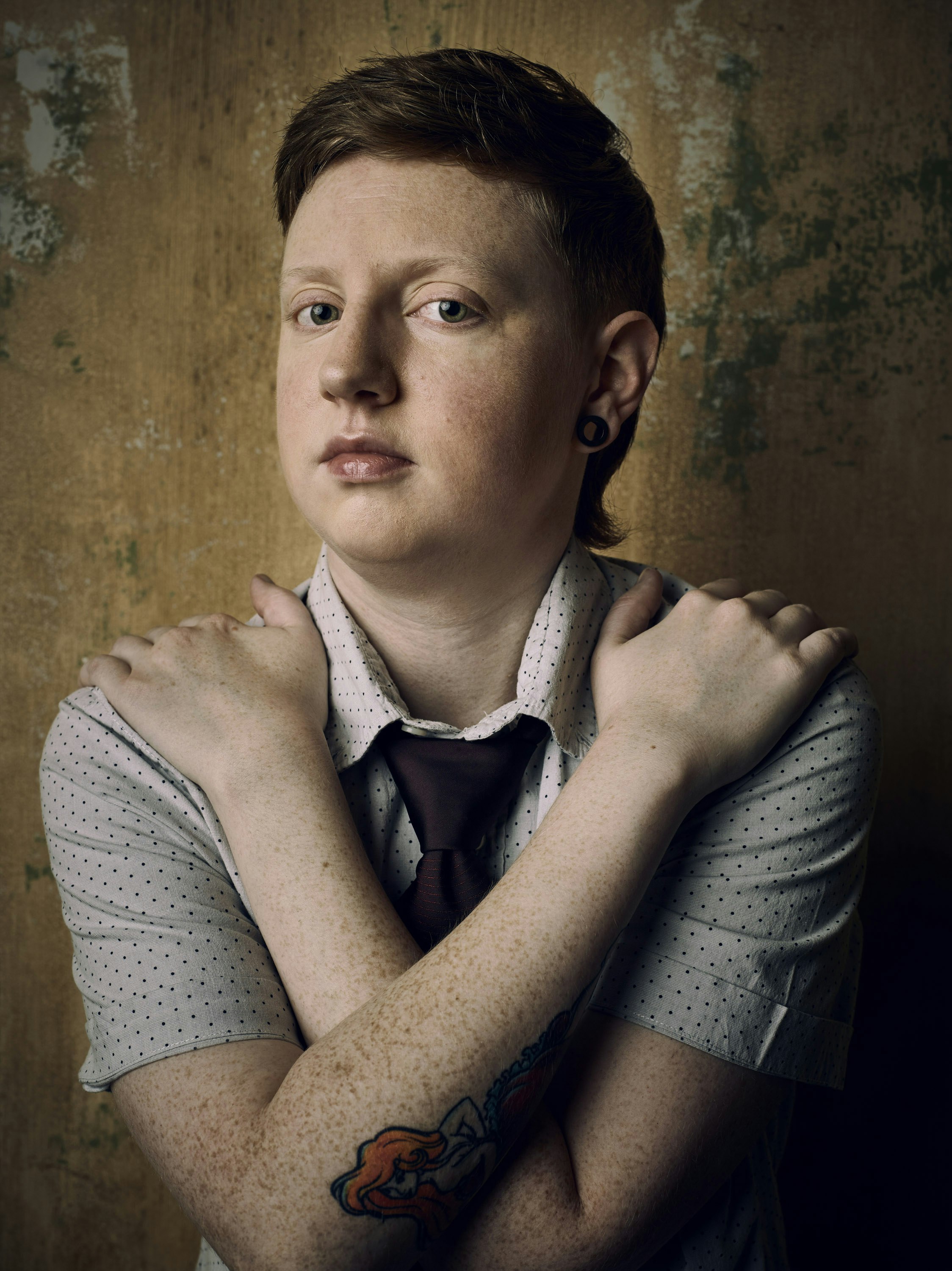 Portrait of a young adult with freckles and short red hair, wearing a white shirt and tie, with crossed arms, against a textured wall.
