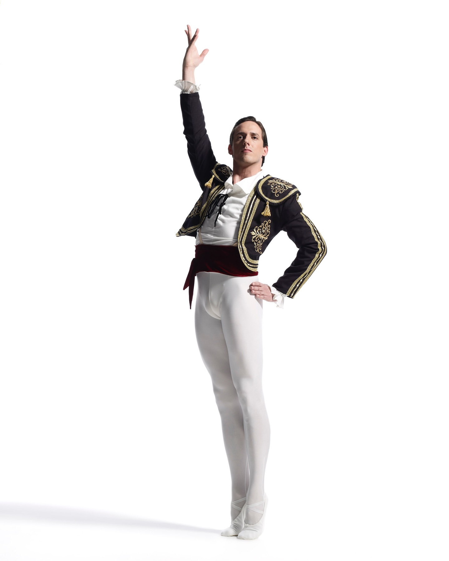 Full-length studio shot of a male ballet dancer in bullfighter-style attire, with one arm raised, set against a stark white backdrop.