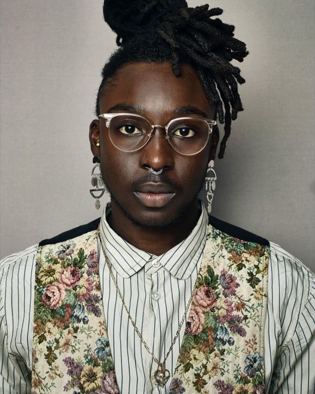 Close-up portrait of a Black adult wearing glasses, a floral vest, striped shirt, earrings, and a nose ring against a grey background.