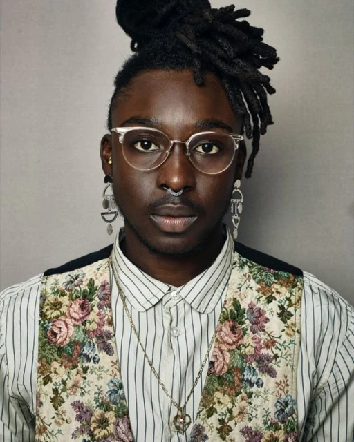 Close-up portrait of a Black adult wearing glasses, a floral vest, striped shirt, earrings, and a nose ring against a grey background.