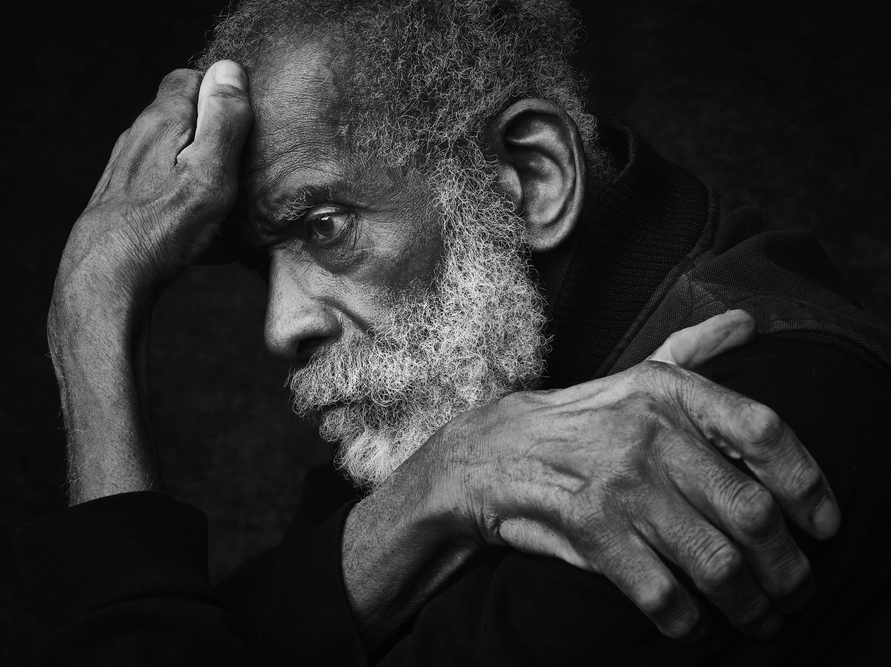 Monochrome portrait of a mature adult man with wrinkled skin and a beard, resting his head in his hands against a dark background.