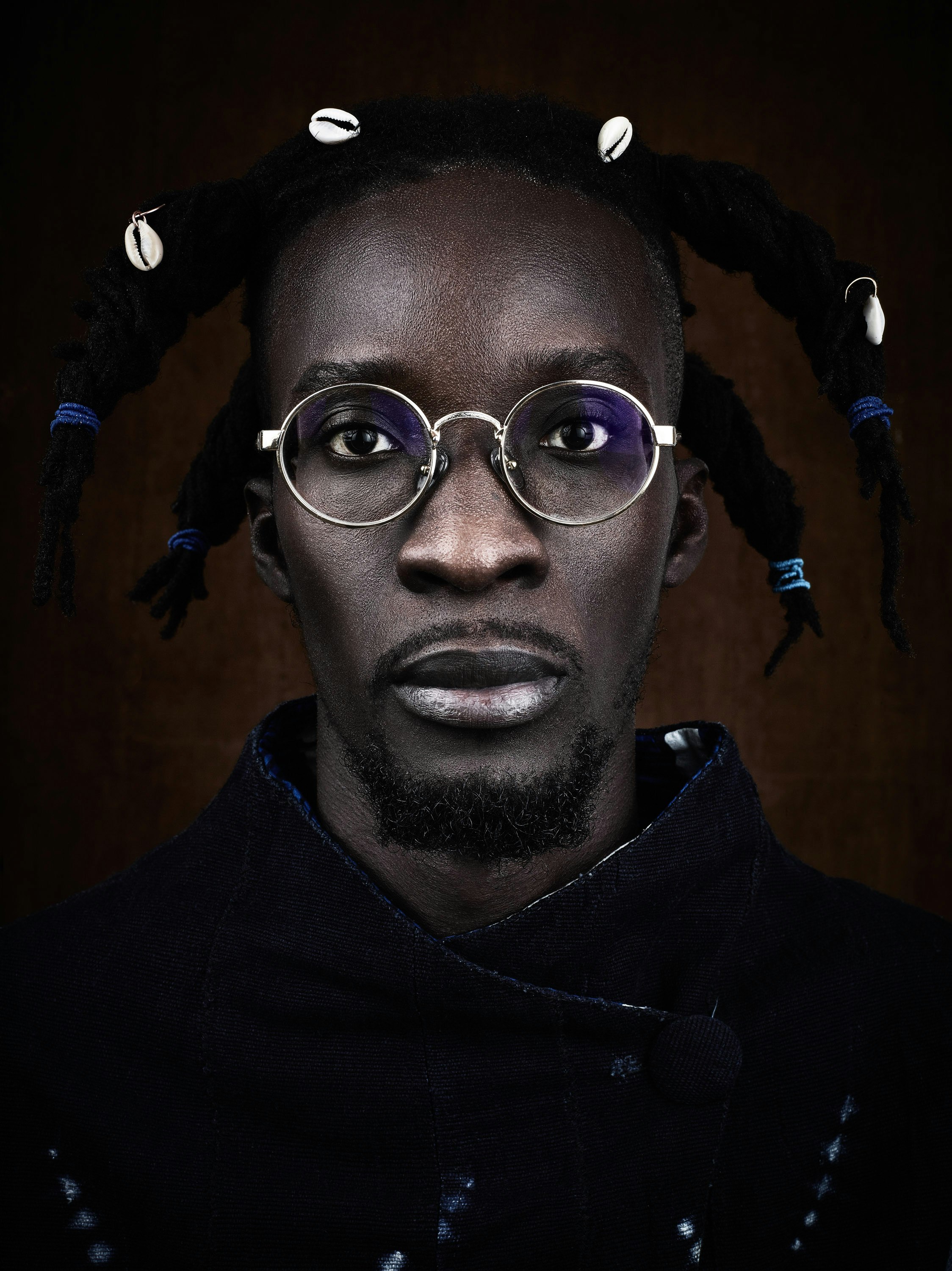 Striking portrait of a Black adult male with dreadlocks adorned with cowrie shells, wearing round glasses and a textured blue jacket.