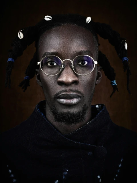 Striking portrait of a Black adult male with dreadlocks adorned with cowrie shells, wearing round glasses and a textured blue jacket.