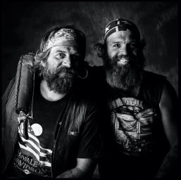 Two bearded men with bandanas and graphic t shirts posing together in a square black and white photographic portrait.