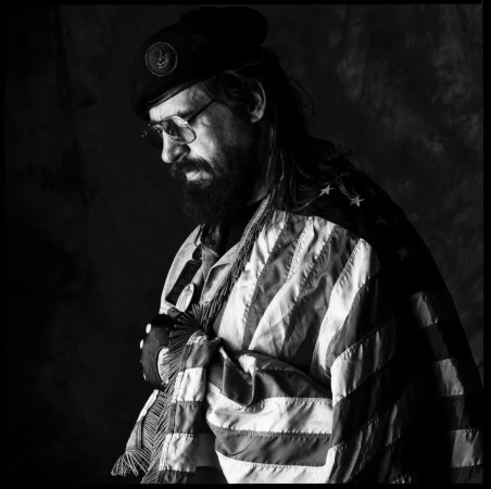Monochrome portrait of a bearded man wearing glasses, a beret, and an American flag draped over his shoulder by Sandro
