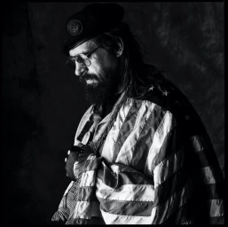 Monochrome portrait of a bearded man wearing glasses, a beret, and an American flag draped over his shoulder by Sandro