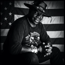 Black man wearing a cowboy hat and fingerless gloves seated against an American flag background in black and white.