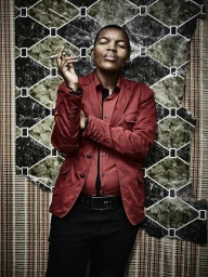Portrait of a person wearing a red blazer and black shirt, holding a cigarette against a patterned wallpaper background.