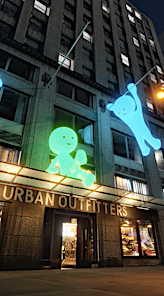 Exterior of Urban Outfitters store with lit-up building decoration art figures on a city building at nighttime, eye-level shot.