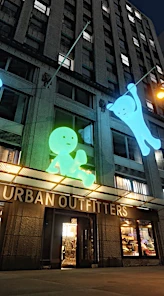 Exterior of Urban Outfitters store with lit-up building decoration art figures on a city building at nighttime, eye-level shot.