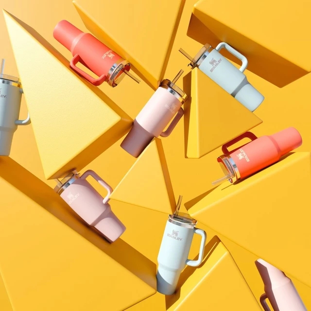 Several Stanley cups in various colors are arranged on a yellow background with geometric shapes, creating a visually appealing product shot.