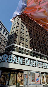 Urban scene featuring a tall building with 'Urban Outfitters' signage, a partially visible pink structure overhead, and clear blue sky.