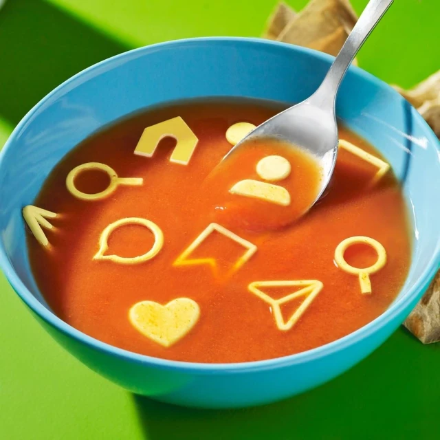 Overhead shot of a bowl of tomato soup with pasta in social media icon shapes, bright lighting, green background, and spoon.
