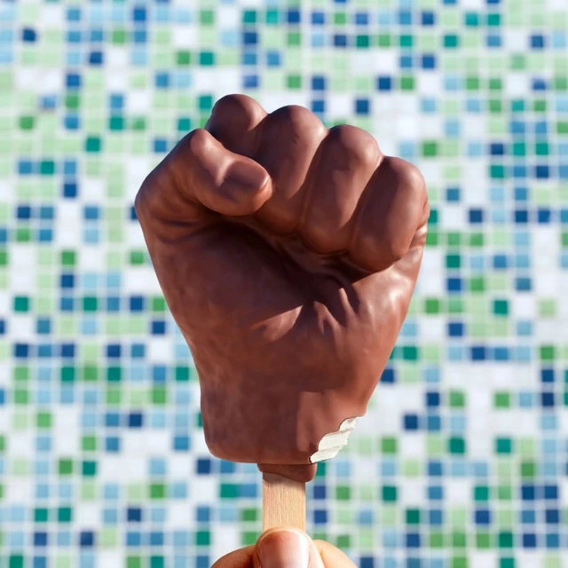 Raised fist ice cream bar held by a hand against a background of blue and green square tile pattern. Confection is partially eaten.