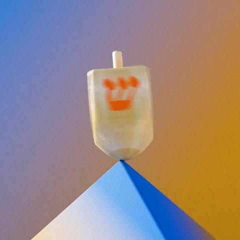 Close-up of a dreidel spinning on a blue geometric base against a purple and yellow background. The dreidel shows blurred Hebrew letters.