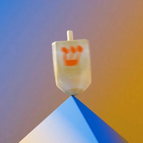 Close-up of a dreidel spinning on a blue geometric base against a purple and yellow background. The dreidel shows blurred Hebrew letters.