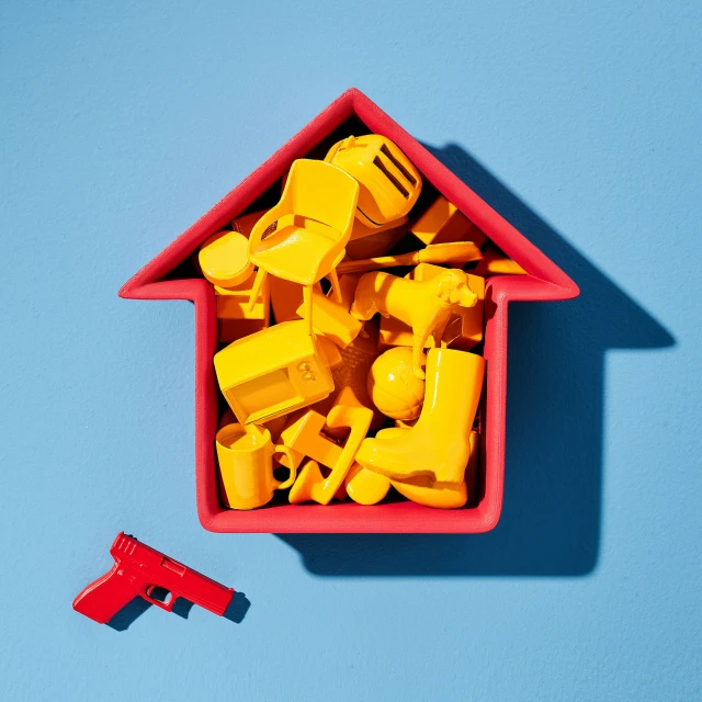 Overhead studio shot of an open-roofed red house shape filled with yellow objects, and a red toy gun beside the house shape, on blue backdrop.