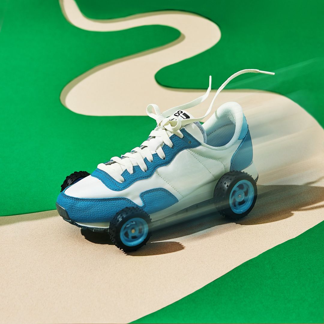 White and blue sneaker with toy wheels pictured on a green and tan background resembling a race track, studio shot.