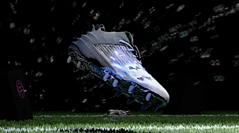 Digitally rendered football cleat in mid-air against a dark background with simulated lights on artificial turf with black box prop.