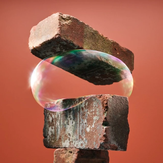 Abstract still life of stacked red bricks supporting a floating iridescent soap bubble against a reddish-brown background.