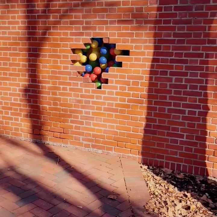 A whimsical red brick wall features a playful hole overflowing with colorful balls on a brick-paved surface with a leaf-strewn edge.