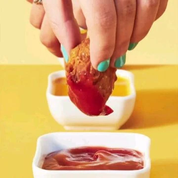 Close-up of a hand dipping a chicken nugget into red sauce, with yellow and red sauces in the background.
