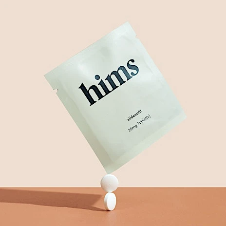 Hims sildenafil tablets in packaging, stacked on white surface against beige backdrop. Medicine and healthcare promotional shot.