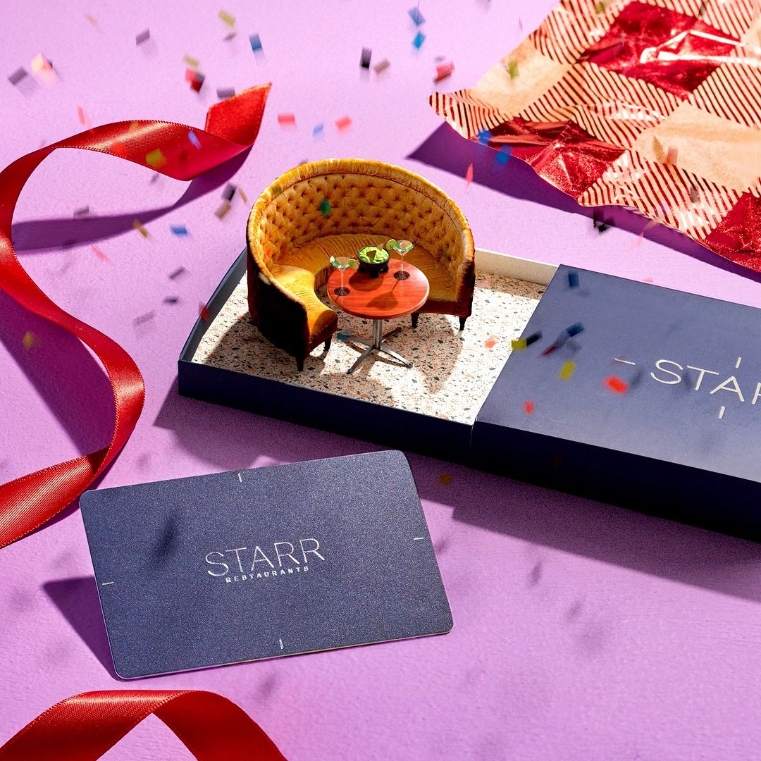 A miniature restaurant scene featuring a small booth, table, and gift card, displayed with confetti and ribbon on a purple background.