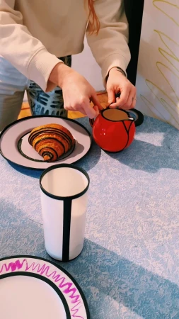 A person arranges a table setting with a croissant, a red pitcher, and a white cup on a blue tablecloth indoors, with a casual, homey vibe.