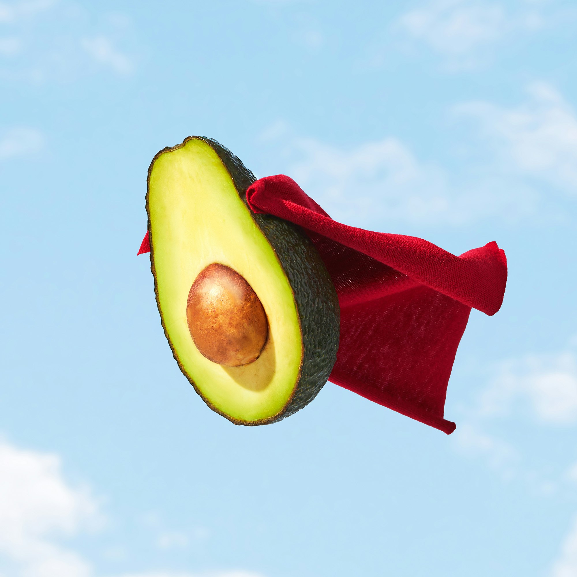 Halved avocado with pit wearing a red cape against a blue sky with soft clouds, suggesting a superhero or healthy food concept.